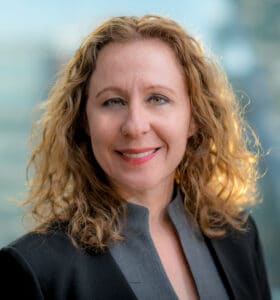 A woman with curly blonde hair smiles at the camera. She is wearing a black blazer over a gray top. The background is blurred, hinting at an outdoor setting.