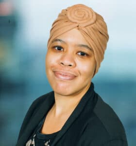 A person wearing a brown headscarf and a black blazer smiles at the camera against a blurred blue background.