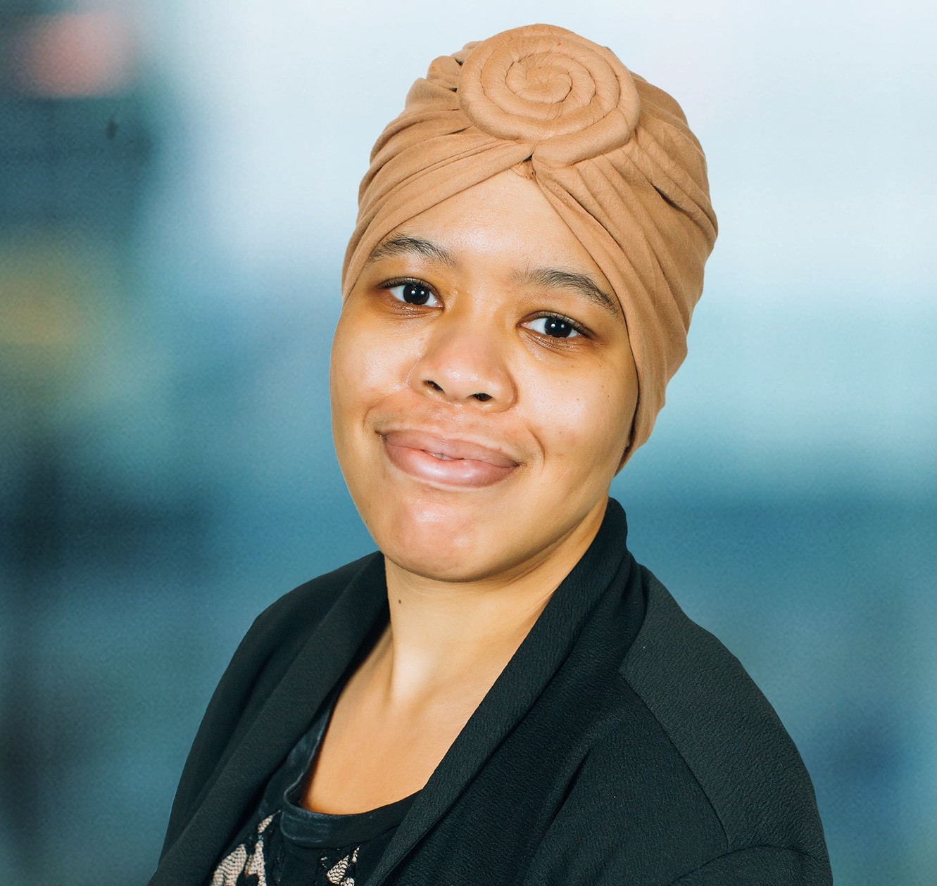 A person wearing a brown headscarf and a black blazer smiles at the camera against a blurred blue background.