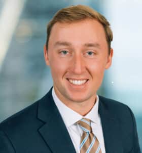 A man in a suit and tie is smiling at the camera. The background is softly blurred, featuring geometric lines and a blue hue.