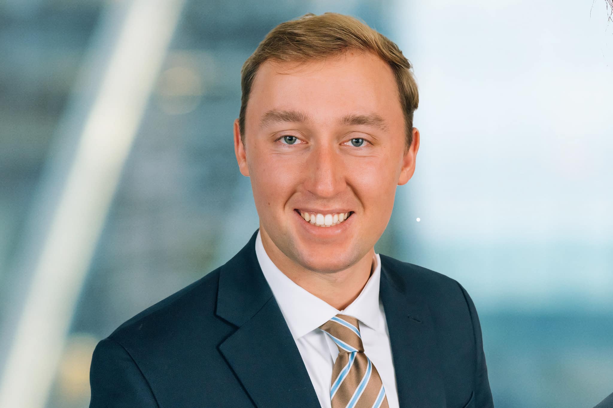 A man in a suit and tie is smiling at the camera. The background is softly blurred, featuring geometric lines and a blue hue.