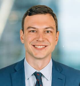 A smiling man in a blue suit and tie stands in front of a blurred office background.