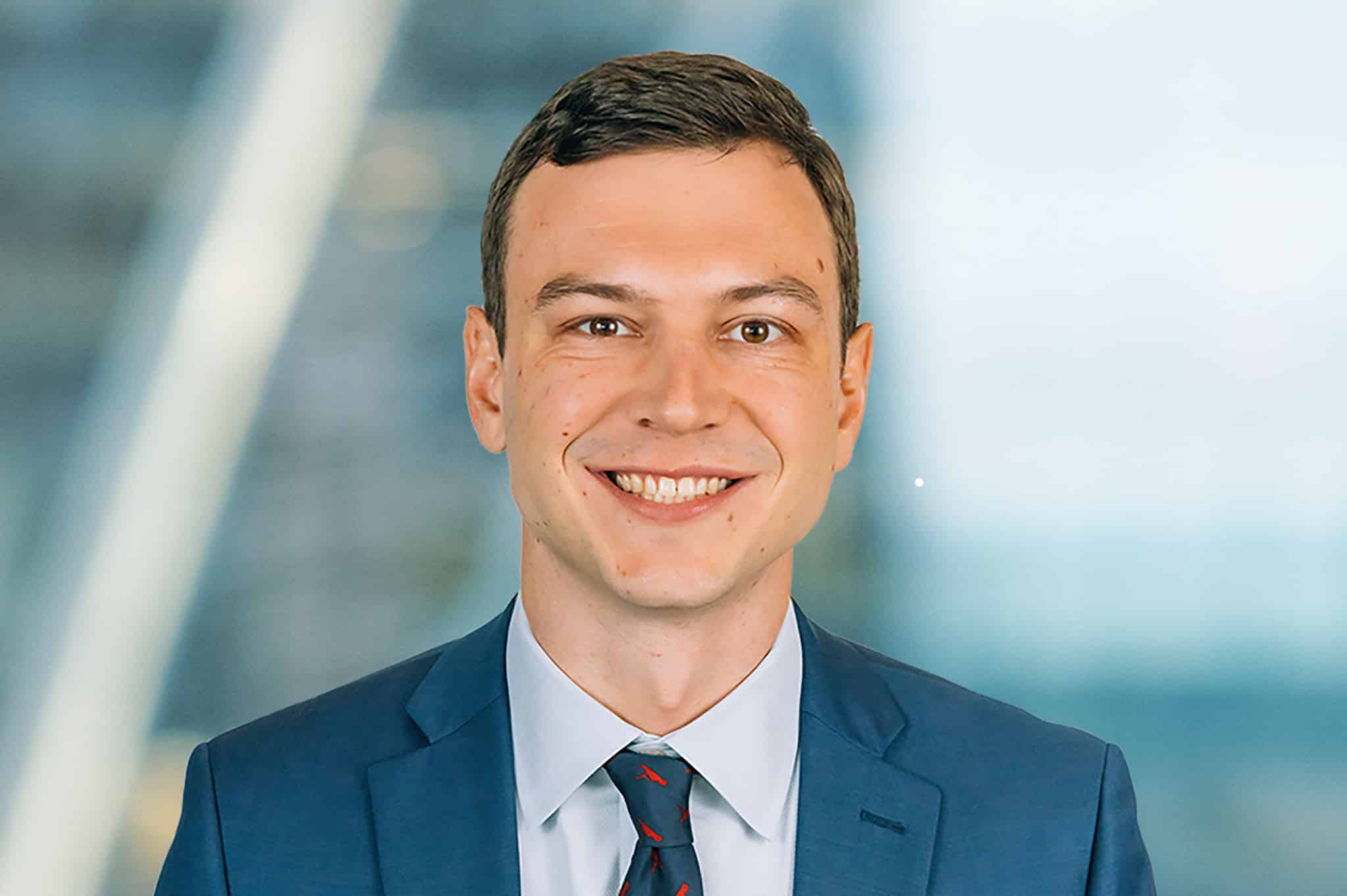 A smiling man in a blue suit and tie stands in front of a blurred office background.