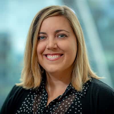 A woman with shoulder-length blonde hair smiling, wearing a black polka-dotted blouse and a black cardigan, with a blurred background.