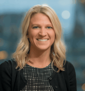 A woman with shoulder-length blonde hair, wearing a black and white patterned top and a black cardigan, smiles at the camera. The background is blurred with soft lighting and out-of-focus buildings.