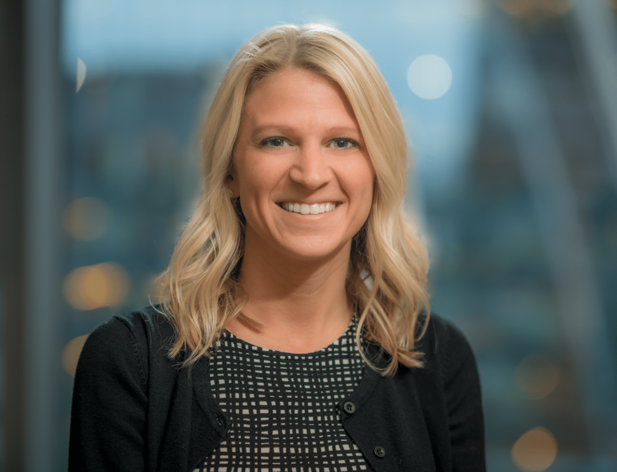 A woman with shoulder-length blonde hair, wearing a black and white patterned top and a black cardigan, smiles at the camera. The background is blurred with soft lighting and out-of-focus buildings.