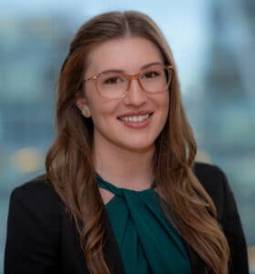 A woman with long brown hair and glasses smiles while wearing a dark blazer and a green blouse. The background is softly blurred, suggesting a cityscape view.