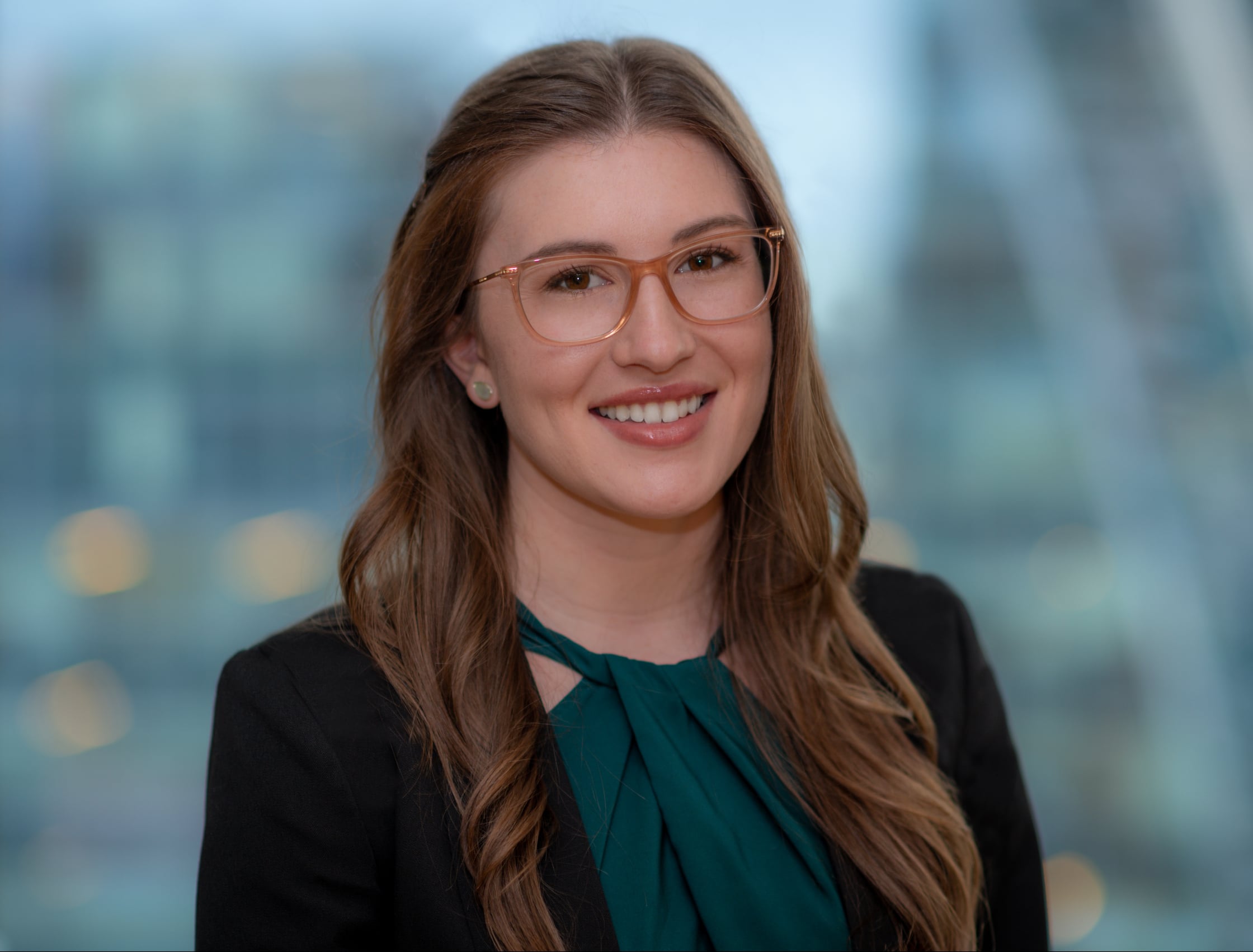 A woman with long brown hair and glasses smiles while wearing a dark blazer and a green blouse. The background is softly blurred, suggesting a cityscape view.