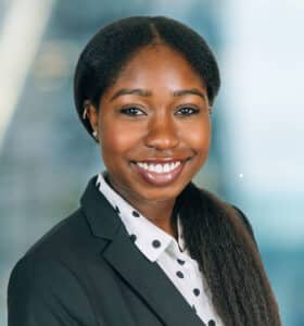 A person wearing a black blazer and polka dot shirt smiles in front of a blurred background with vertical and diagonal lines.