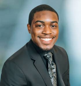 A person in a suit sits against a blue background. They are smiling and wearing a dark jacket, a tie with a paisley pattern, and a black shirt.