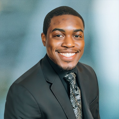 A person in a suit sits against a blue background. They are smiling and wearing a dark jacket, a tie with a paisley pattern, and a black shirt.