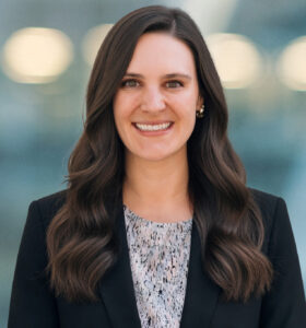 A woman with long brown wavy hair, wearing a black blazer and a patterned blouse, smiles at the camera with a blurred background of lights and soft blue tones.