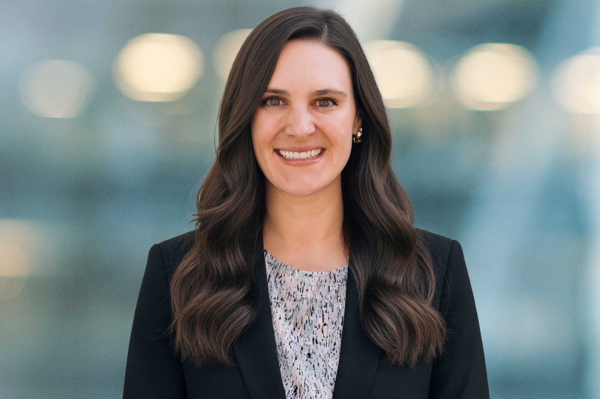 A woman with long brown wavy hair, wearing a black blazer and a patterned blouse, smiles at the camera with a blurred background of lights and soft blue tones.