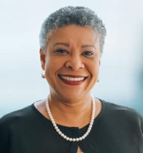 Smiling person with short gray hair wearing a black top and pearl necklace against a blurred background.