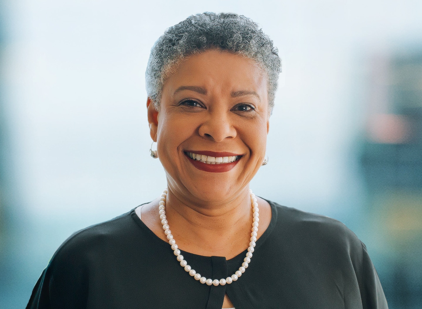 Smiling person with short gray hair wearing a black top and pearl necklace against a blurred background.