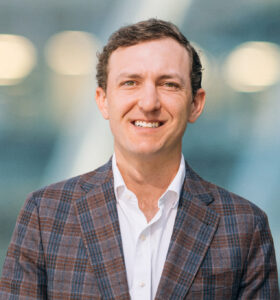 A man with short brown hair, wearing a checked blazer and white shirt, smiles at the camera. The background is blurred with blue and white tones and circular lights.