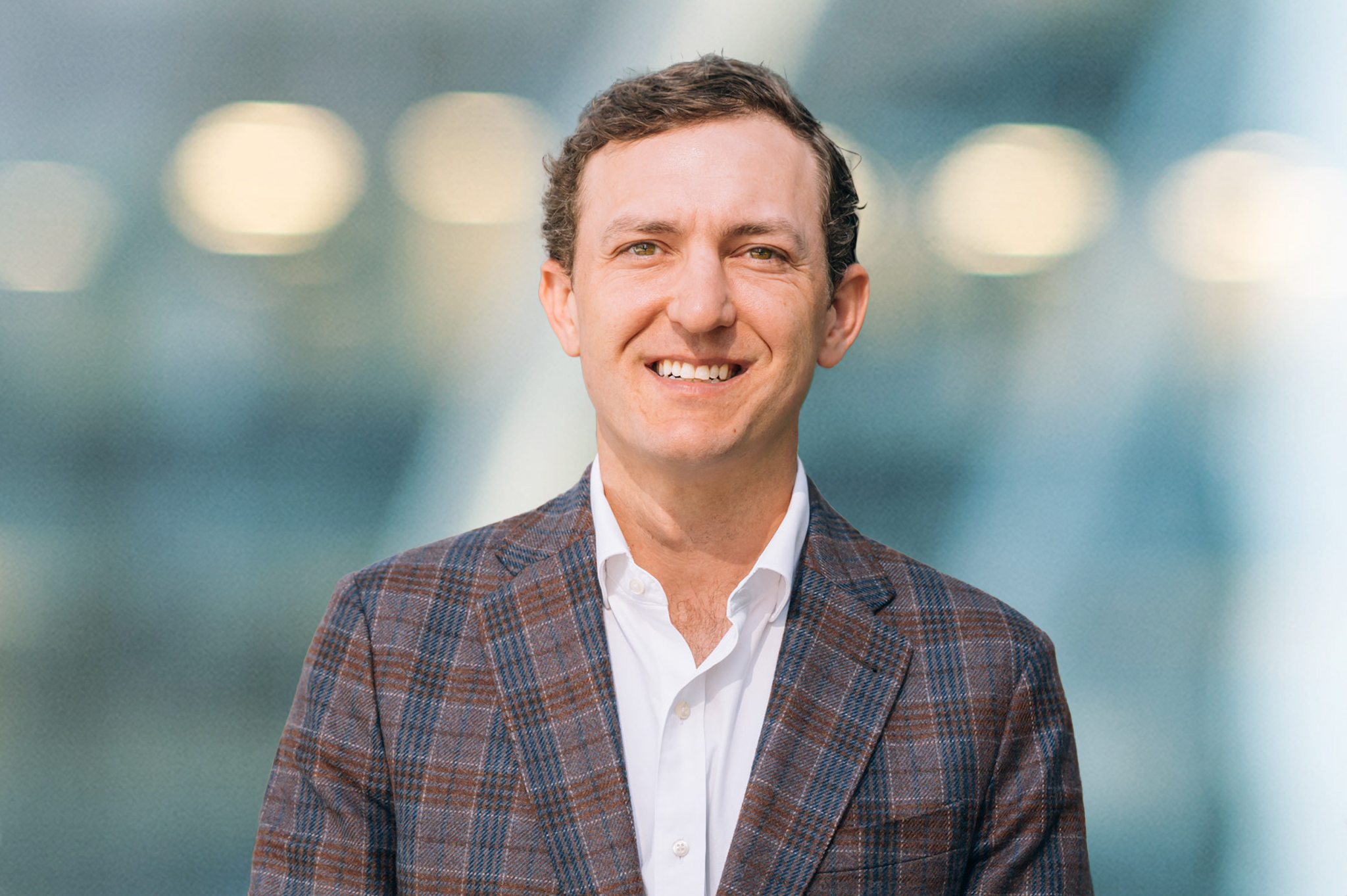 A man with short brown hair, wearing a checked blazer and white shirt, smiles at the camera. The background is blurred with blue and white tones and circular lights.