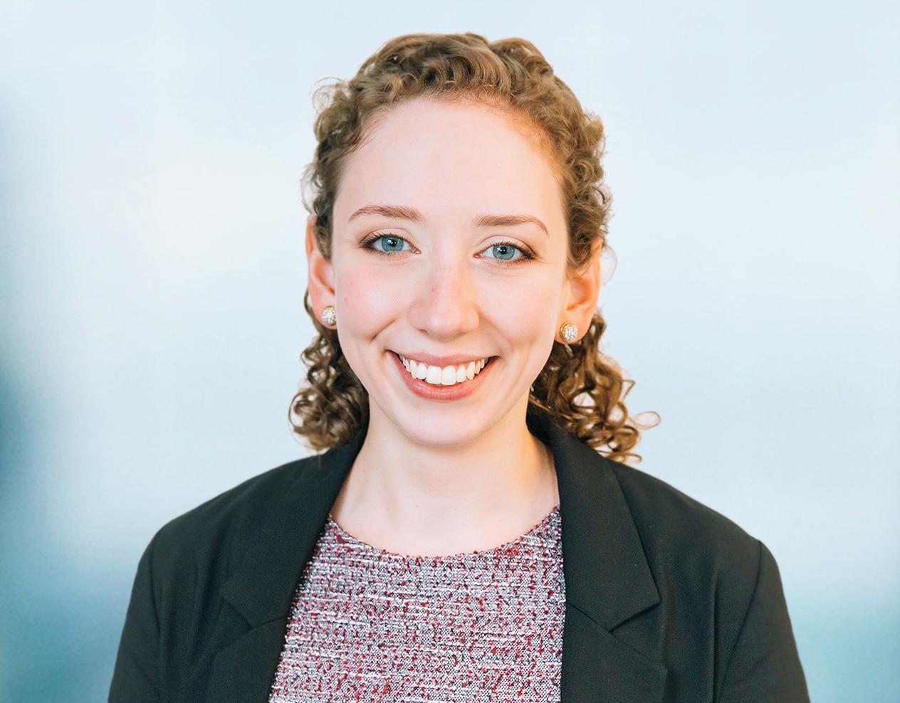 A person with curly hair is smiling while wearing a black blazer and a patterned top against a light blue background.