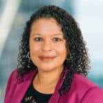 A person with curly hair is smiling, wearing a magenta blazer over a black top. The background is softly blurred, suggesting an indoor setting.
