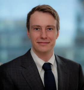 A young man in a suit and tie is smiling against a blurred cityscape background. He has short brown hair and is wearing a checkered jacket with a white shirt.