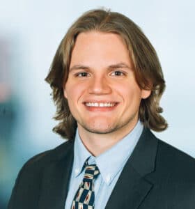 A person with shoulder-length hair is smiling, wearing a suit, a light blue shirt, and a patterned tie. The background is out of focus.