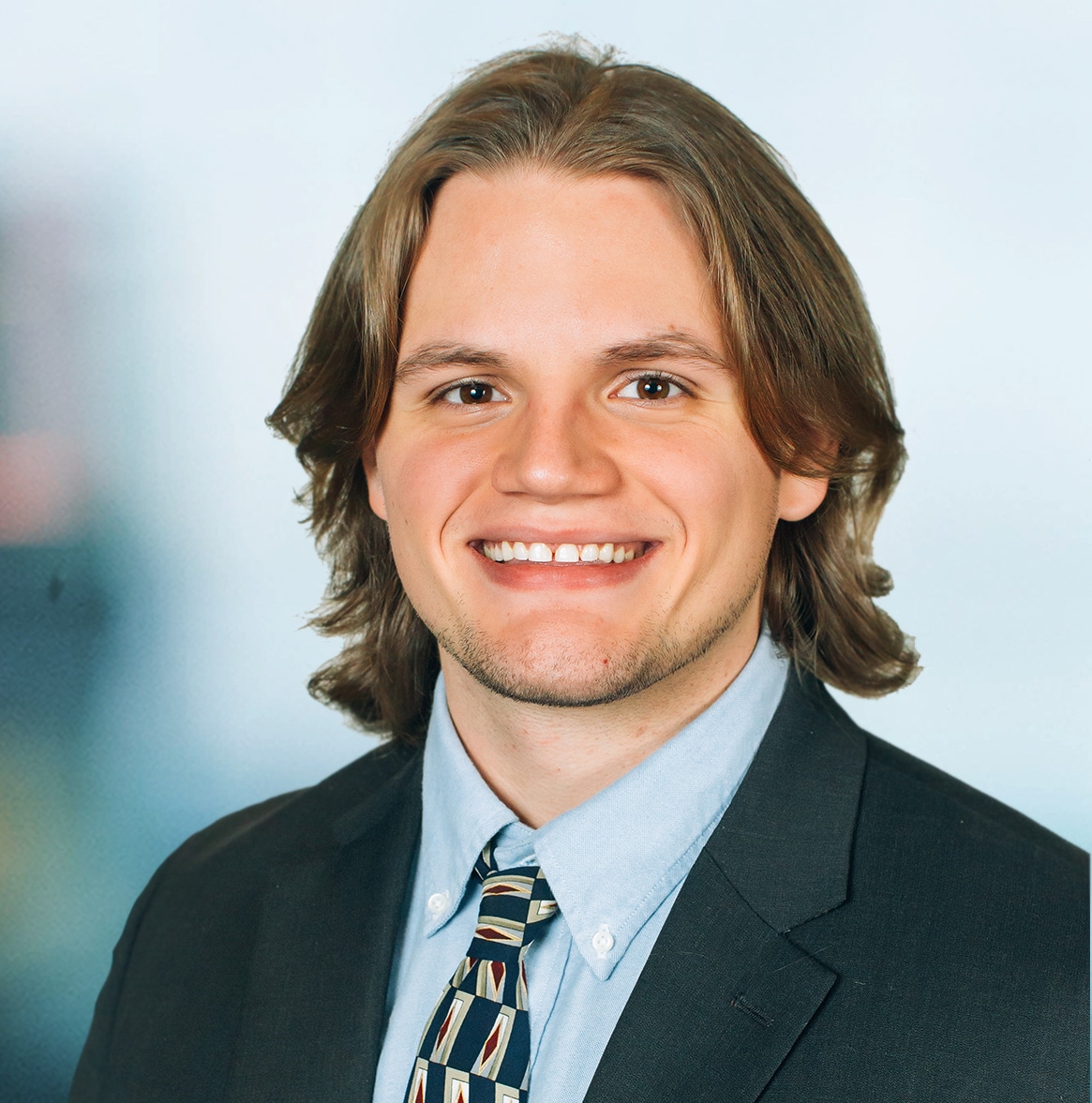 A person with shoulder-length hair is smiling, wearing a suit, a light blue shirt, and a patterned tie. The background is out of focus.