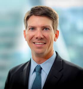 A man in a suit and tie smiles at the camera. The background is blurred, showing a mix of blue and gray tones.