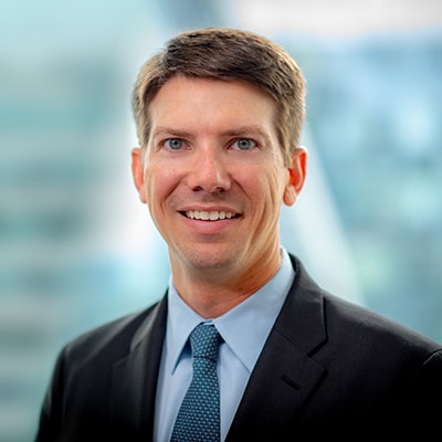 A man in a suit and tie smiles at the camera. The background is blurred, showing a mix of blue and gray tones.