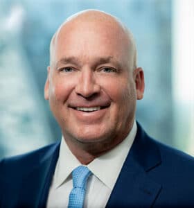 A man in a blue suit and light blue tie is smiling in front of a blurred background.