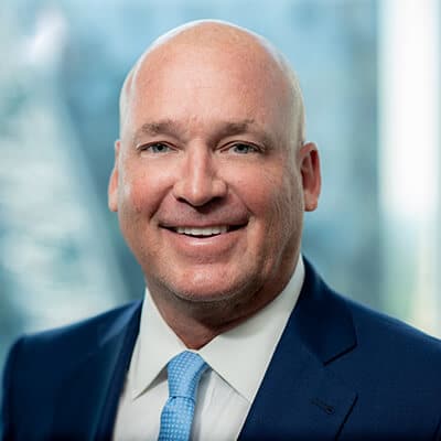 A man in a blue suit and light blue tie is smiling in front of a blurred background.