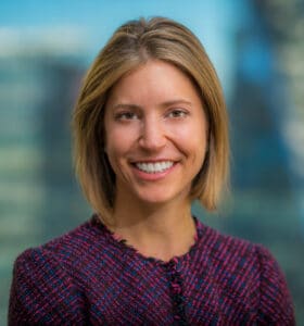 A smiling person with short, blond hair is wearing a textured, dark-colored jacket. The background is blurred, showing an abstract urban setting with hints of glass buildings.