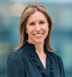 A woman with straight, shoulder-length light brown hair is smiling, wearing a black blouse with ruffle details, standing in front of a blurred blue background.