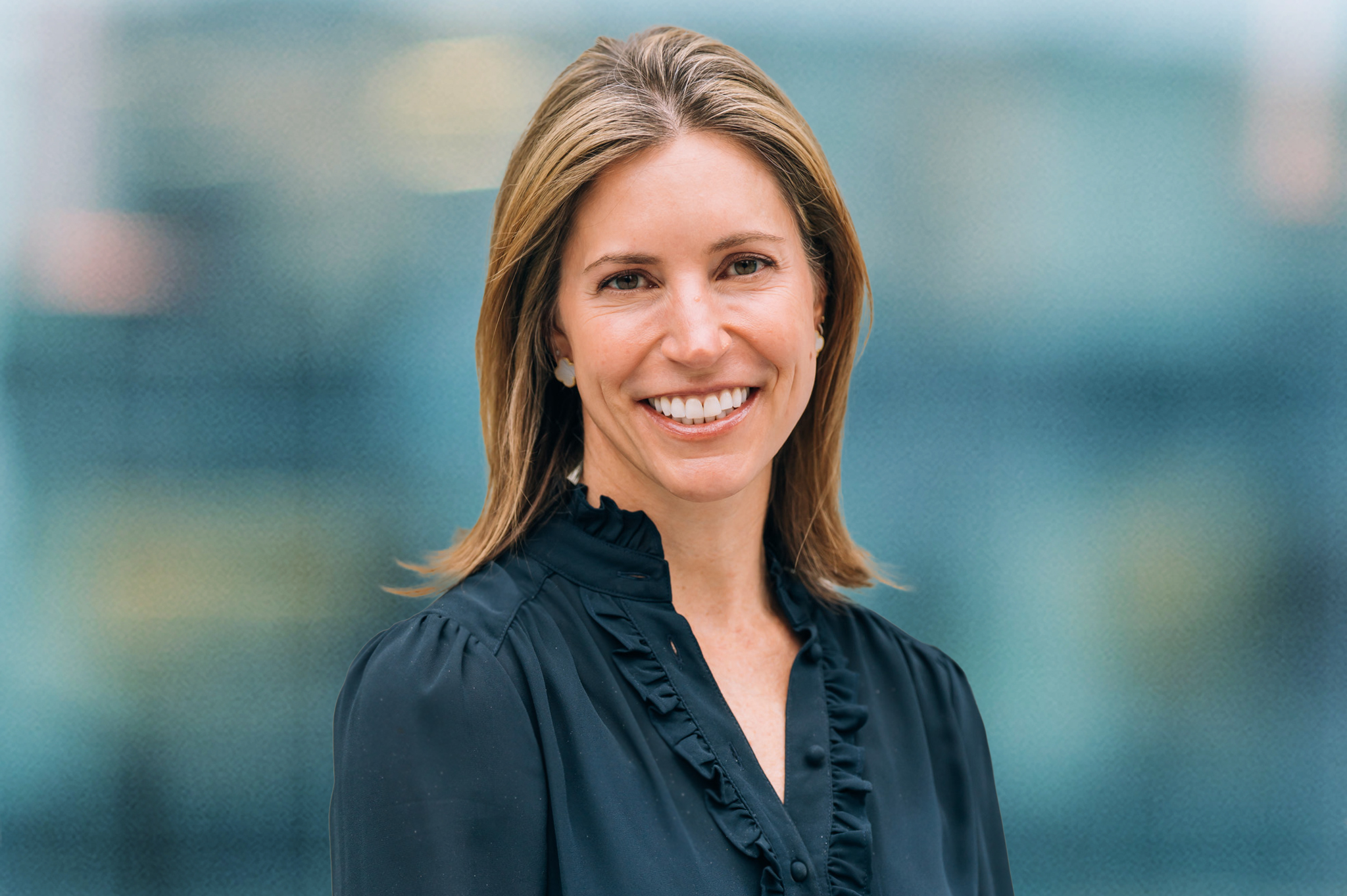 A woman with straight, shoulder-length light brown hair is smiling, wearing a black blouse with ruffle details, standing in front of a blurred blue background.