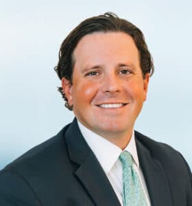 A smiling man with short, wavy brown hair is wearing a dark suit, white shirt, and light blue patterned tie. He is sitting against a light blue background.