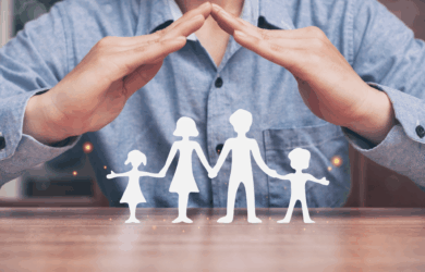 A person in a blue shirt creates a protective gesture with their hands above a cut-out silhouette of a family with two adults and two children on a wooden table.