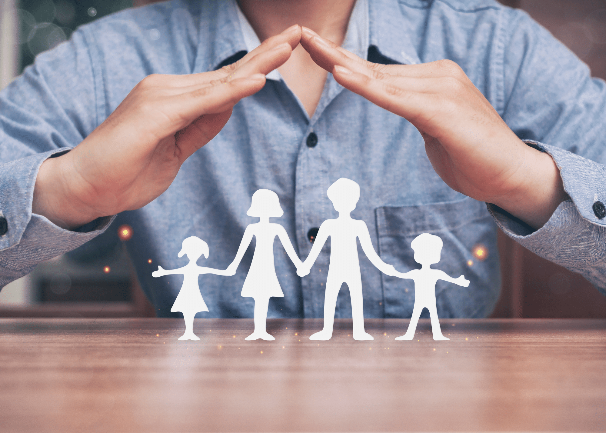 A person in a blue shirt creates a protective gesture with their hands above a cut-out silhouette of a family with two adults and two children on a wooden table.
