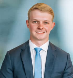A man with short blonde hair is wearing a dark suit, white shirt, and light blue tie. He is smiling and positioned against a blurred blue-gray background.