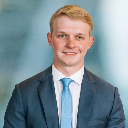 A man with short blonde hair is wearing a dark suit, white shirt, and light blue tie. He is smiling and positioned against a blurred blue-gray background.