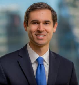A man in a dark suit and blue tie is smiling in a professional setting with a blurred cityscape in the background.