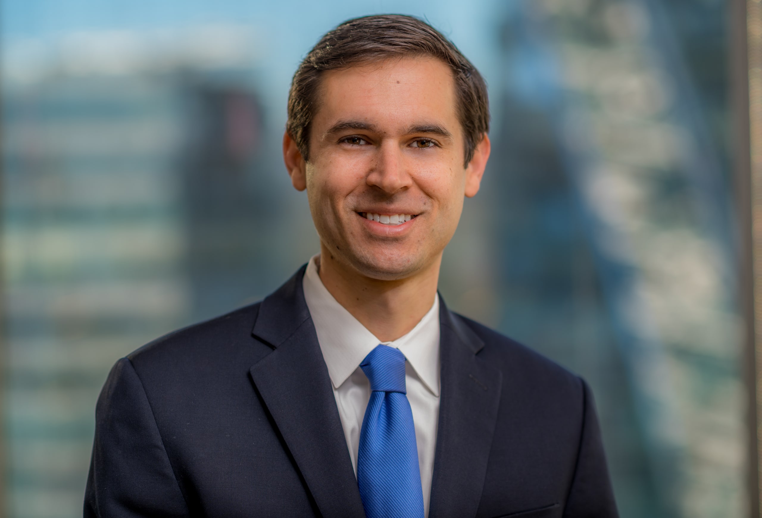 A man in a dark suit and blue tie is smiling in a professional setting with a blurred cityscape in the background.