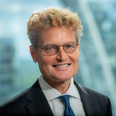 Frank-Butterfield A man with curly blonde hair and glasses wearing a dark suit, white shirt, and blue tie, smiling against a blurred background.