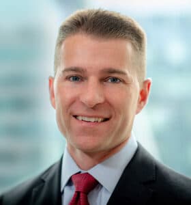 A person in a suit and red tie smiles at the camera. The background is blurred, suggesting an office setting.
