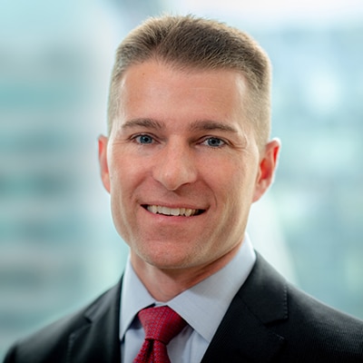 A person in a suit and red tie smiles at the camera. The background is blurred, suggesting an office setting.