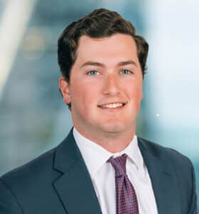 A smiling person with short, dark hair wearing a suit, white shirt, and red patterned tie. The background is blurred with a mix of blue and light colors.