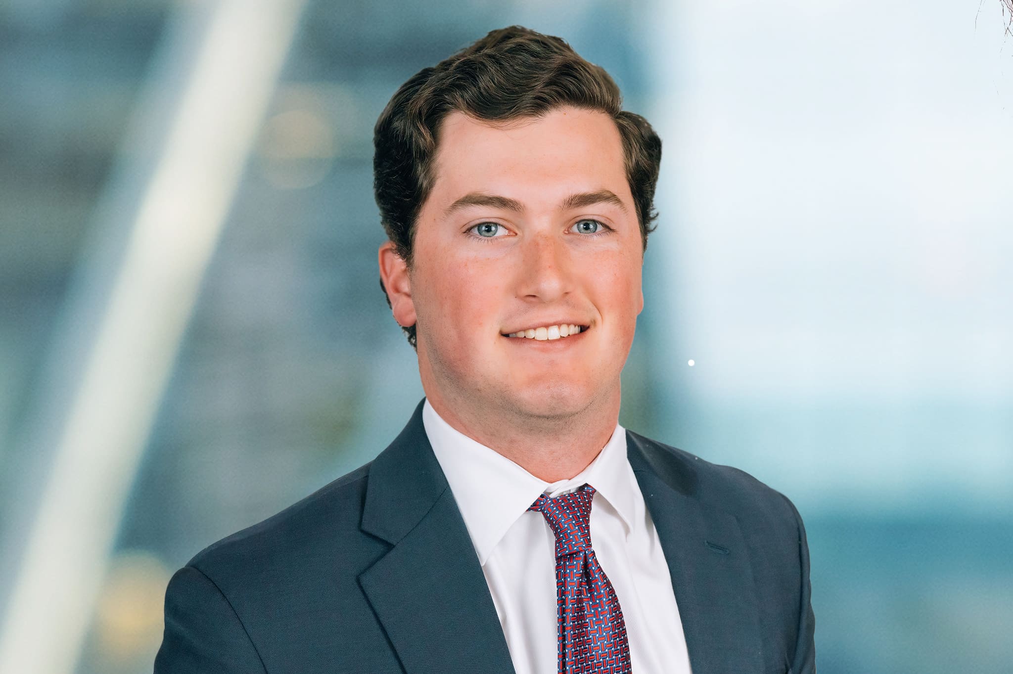 A smiling person with short, dark hair wearing a suit, white shirt, and red patterned tie. The background is blurred with a mix of blue and light colors.