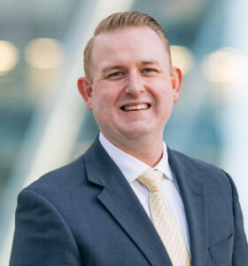 A man in a gray suit and light yellow tie smiles at the camera. The background is blurred with soft blue and white tones and round lights, suggesting an outdoor or modern office setting.