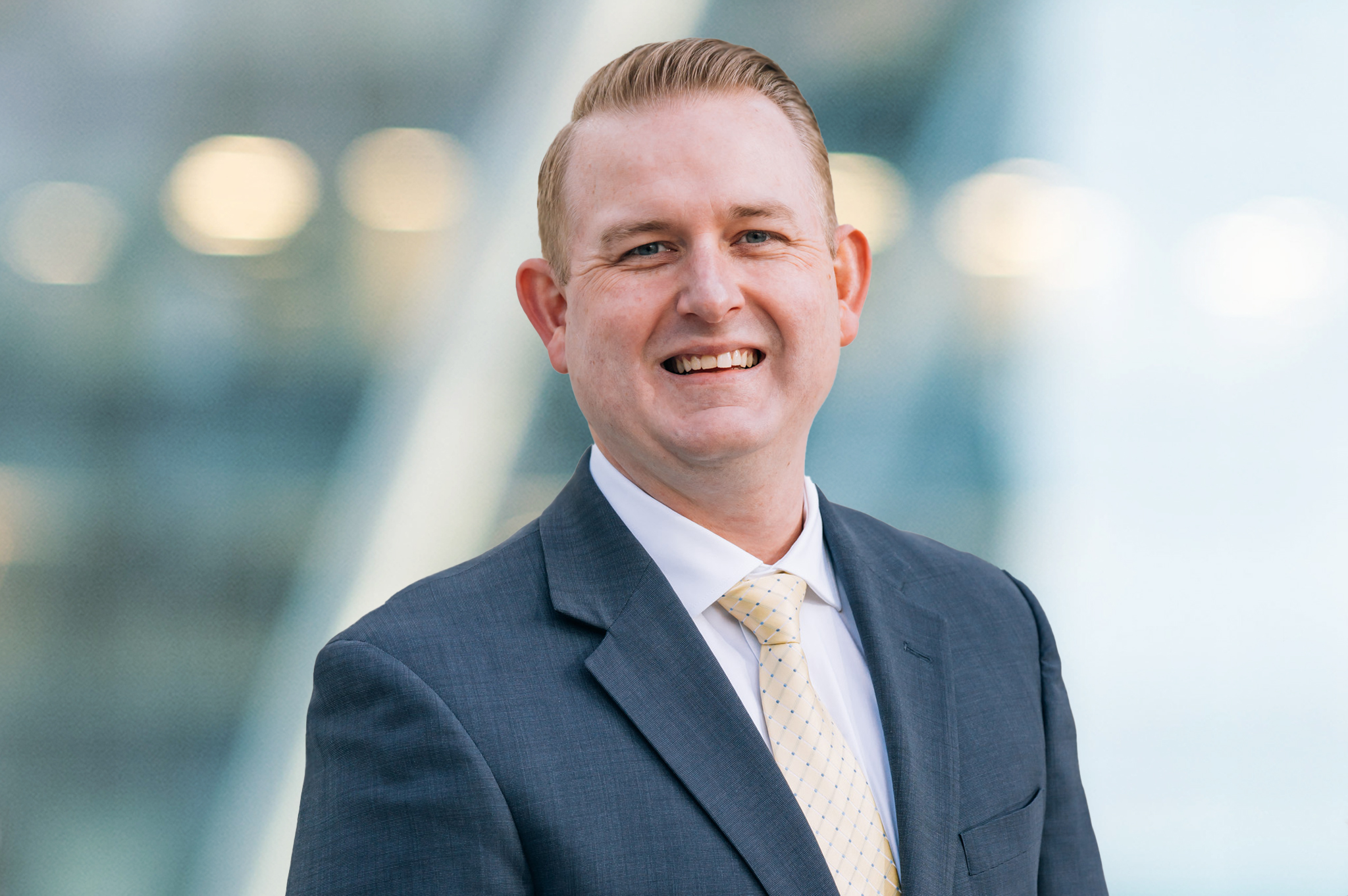 A man in a gray suit and light yellow tie smiles at the camera. The background is blurred with soft blue and white tones and round lights, suggesting an outdoor or modern office setting.