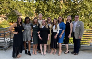 A group of ten adults smiling and standing together outdoors on a patio with trees in the background. They are dressed in formal attire, and some are wearing name tags.
