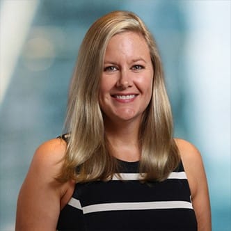 A person with long blonde hair smiles at the camera. They are wearing a black and white striped sleeveless top. The background is softly blurred.