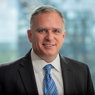 A man wearing a dark suit, white shirt, and blue striped tie is smiling in front of a blurred, modern office building background.